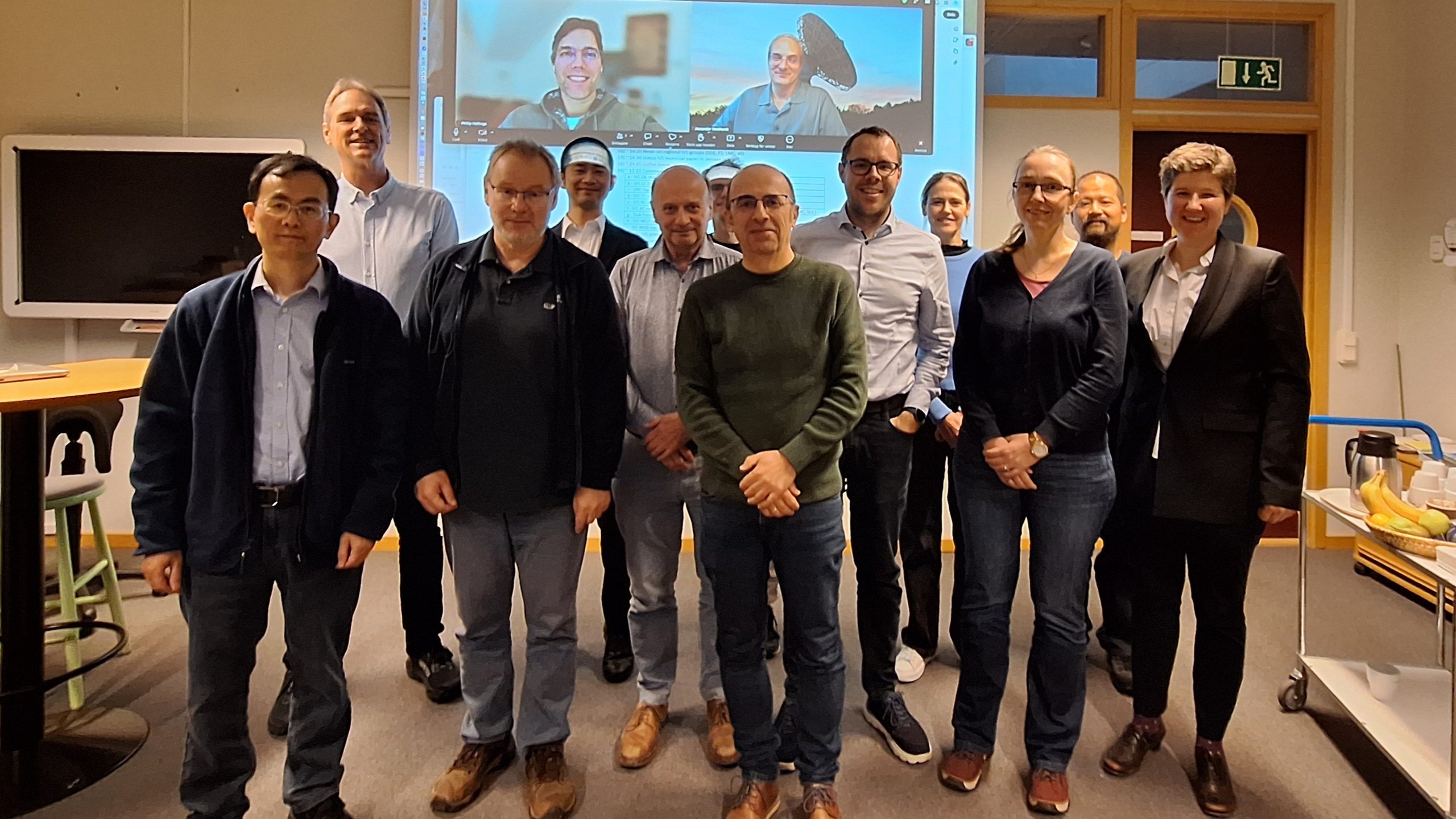 A group photo of twelve people standing and smiling at the camera, with two people on the projector screen behind them, also smiling at the camera